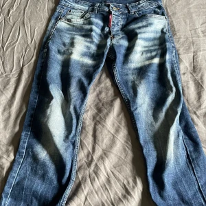 Blå Dsquared2 jeans  - Säljer ett par blå jeans från Dsquared2 med coola slitningar och ljusa tvättade partier. Klassisk femficksmodell med röd logotyp vid gylfen och snygga kontrastsömmar. Jeansen har rak passform och är tillverkade i bomullsdenim.