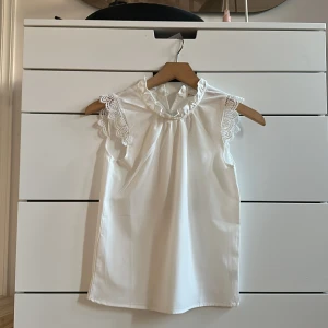 Vit blus med spetsdetaljer - Säljer en söt vit blus med volangkrage och spetsdetaljer på ärmarna. Blusen är ärmlös och har en rak passform. 