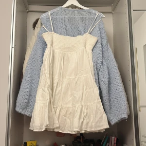 Vit kort sommarklänning med smock - Supersöt vit klänning med smala axelband och smock upptill. Kjolen är volangig och luftig, perfekt för varma dagar. Klänningen är kort och har en romantisk vibe med sin flowiga passform. Materialet känns lätt och skönt mot huden.