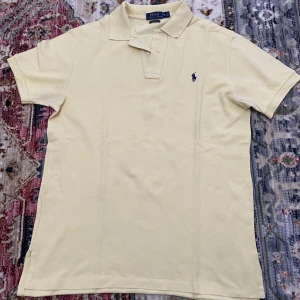 Pikétröja från Polo Ralph Lauren - Snygg ljusgul pikétröja från Polo Ralph Lauren i klassisk passform. Tröjan har korta ärmar, krage och två knappar vid halsen. Ikoniska Polo-loggan i mörkblått broderad på bröstet. 