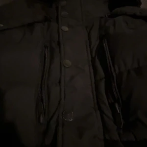 Svart pufferjacka från Smog, S - Svart pufferjacka från Smog i storlek S. Jackan har dragkedjor på bröstfickorna, tryckknappar framtill och en hög krage. Perfekt för kalla dagar med sin vadderade design och stilrena look. PRIS KAN DUSKITERAS 