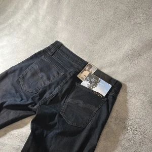 Svarta jeans från Nudie Jeans (helt nya) - Snygga svarta skinny jeans från Nudie Jeans med klassisk femficksdesign och diskret broderad bakficka. Jeansen har smal passform och är tillverkade i stretchig bomullsdenim för en bekväm känsla. Perfekta för dig som gillar en stilren look. Model skinny lin. W29 L 34