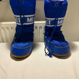 Blå Moon Boot snowboots - Säljer ett par klassiska blå Moon Boot snowboots med vit logga och snörning framtill. Skorna har en hög modell med rund tå och platt sula i gummi, perfekt för vintern. Yttermaterialet är syntet och de är fodrade för extra värme. Passar 42