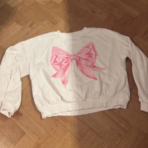Vit sweatshirt med rosa rosett Lindex - Vit sweatshirt från Lindex med en stor rosa rosett framtill. Tröjan har rund hals, långa ärmar och ribbade muddar vid ärmslut och nederkant. Tillverkad i mjuk bomullsmix, perfekt för en söt och avslappnad stil.