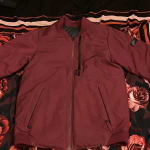 Vinröd bomberjacka från Nike - Säljer en vinröd bomberjacka från Nike med quiltat svart foder. Jackan har dragkedja framtill, två snedställda fickor och en bröstficka med dragkedja. Ribbade muddar vid krage, ärmslut och nederkant. Materialet är polyester.