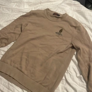Beige stickad tröja från Morris strl M - Snygg beige stickad tröja från Morris med broderad logga och emblem på bröstet. Tröjan har rund halsringning, ribbade muddar och är långärmad. Perfekt för dig som gillar klassisk och stilren look.