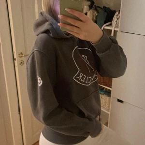 Grå hoodie från Moncler med stor logga - Säljer en grå hoodie från Moncler med stor broderad logga i vitt och rött på bröstet. Tröjan har huva med snörning, känguruficka och ribbade muddar. Klassisk streetwear-look och perfekt för dig som gillar exklusiva märken.
