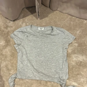 Grå kortärmad topp från Kappahl - Säljer en ljusgrå kortärmad topp från Kappahl i mjuk bomullsmix. Toppen har rund halsringning och knyt-detalj i nederkant som ger en avslappnad och trendig look. Perfekt till jeans eller shorts för en chill stil.