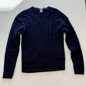 Mörkblå stickad tröja med kabelmönster - Mysig mörkblå stickad tröja från Lindex med klassiskt kabelstickat mönster. Tröjan har rund halsringning och långa ärmar, perfekt för kyligare dagar. Ribbstickade muddar vid ärmslut och nederkant ger en snygg finish.