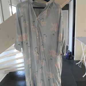Ljusblå blommig långklänning - Säljer en ljusblå långklänning med kaprifol mönster i rosa och grönt. Klänningen har knappar framtill, trekvartsärm och en avslappnad passform. Perfekt för dig som gillar en chill och feminin stil.
