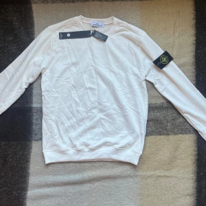 Vit sweatshirt från Stone Island - Säljer en clean vit sweatshirt från Stone Island med klassisk logga på vänster ärm. Tröjan har rund hals, långa ärmar och är gjord i mjuk bomull. Perfekt för dig som gillar stilrena och exklusiva plagg med streetkänsla.
