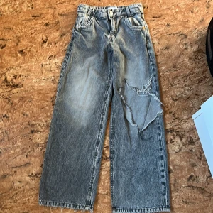Gråa wide jeans från Zara med slitningar - Säljer ett par gråa jeans från Zara i modell wide med rejäl slitning och trasiga detaljer på ena benet. Jeansen har råa, fransiga benslut och klassisk femficksdesign. Perfekta för dig som gillar en edgy och avslappnad stil.