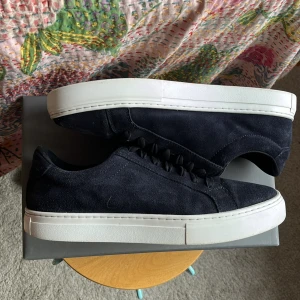 Mörkblå mockasneakers från Vagabond - Snygga mörkblå sneakers från Vagabond i mjuk mocka med vita, platta gummisulor. Klassisk låg modell med rund tå och mörkblå snören. Perfekta för dig som gillar stilrena och minimalistiska skor med en modern touch. Skornas skick är 6/10 mockan är lite mer sliten vid tårarna och där byxorna skaver, nypris 1400 mitt pris 600 man får även med original boxen, hör av er vid mer frågor.