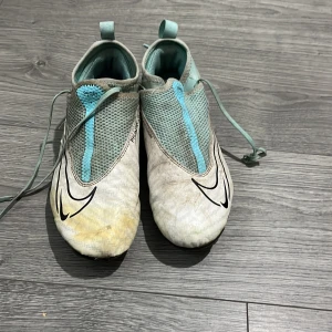 Nike Phantom Vision fotbollsskor ljusblå - Nike Phantom Vision fotbollsskor i ljusblått och vitt med svarta detaljer. Skorna har snörning, meshpanel upptill och klassisk Nike Swoosh på sidan. Yttersulan har fasta dobbar för bra grepp på planen. Perfekta för dig som vill sticka ut på fotbollsplanen.