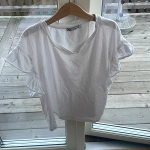 Vit blus med volangärm från Zara - Säljer en vit blus från Zara med korta volangärmar och avslappnad passform. Toppen är tillverkad i mjuk bomull, perfekt för en fräsch och enkel look.