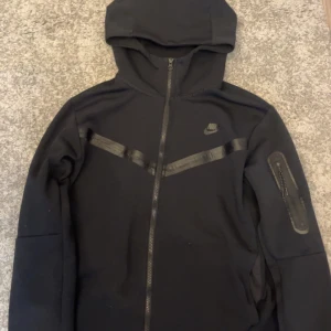 Svart Nike hoodie med dragkedja - Svart hoodie från Nike med hel dragkedja framtill och huva. Snygga svarta detaljer över bröstet och en extra ficka med dragkedja på ärmen. Perfekt för dig som gillar sportig och clean stil. Nike-logga på bröstet och mjukt material.