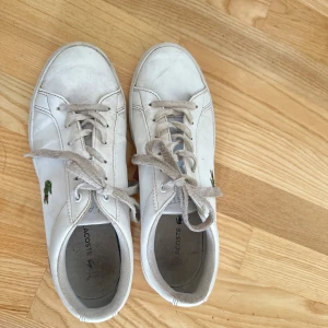 Vita sneakers från Lacoste - Klassiska vita sneakers från Lacoste med lågt skaft och snörning. Skorna har den ikoniska gröna krokodilen broderad på sidan och är tillverkade i skinn. Perfekta för en clean och stilren look.