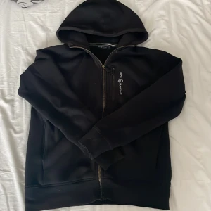 Svart hoodie från Sail Racing - Svart hoodie från Sail Racing med dragkedja framtill och huva. Tröjan har en diskret logga på bröstet och praktiska fickor på sidan. Tröjan har ett litet hål vid ena ärmen men inget som syns jättemycket men därav priset. Pris kan diskuteras, hör av dig vid minsta fundering!