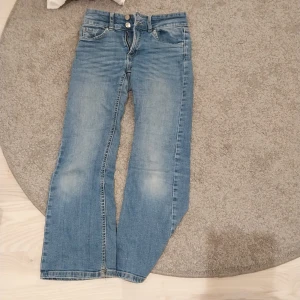 Blå bootcut jeans från Denim Day - Snygga blå jeans från Denim Day i storlek 140. Modellen har bootcut-ben, klassiska fickor fram och bak med snygga detaljer och knappar. Jeansen är i mjuk denim med lätt slitning för en cool look.