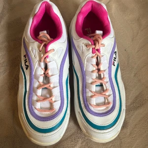 Färgglada Fila sneakers med pastell - Säljer ett par chunky Fila sneakers i vitt med detaljer i lila, turkos och rosa. Skorna har rosa snören, vit sula och en cool retrovibe. Insidan är fodrad i stark rosa och materialet är en mix av syntet och textil. Perfekta för dig som gillar färg och streetstyle.