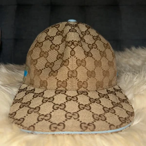 Gucci keps med monogram och blå detalj - Säljer en beige och brun keps från Gucci med klassiskt GG-monogram över hela kepsen. Kepsen har en ljusblå justerbar rem bak och en beige/blå rand på sidan. Materialet är bomullscanvas och insidan är brun. Perfekt för dig som vill sticka ut med lyxig streetstyle.