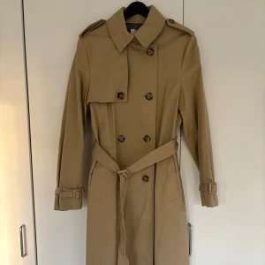 Beige trenchcoat från Mango, stl M - Klassisk beige trenchcoat från Mango med dubbelknäppning, axelklaffar och bälte i midjan. Jackan har långa ärmar med spännen vid handlederna och två fickor framtill. Perfekt för dig som vill ha en tidlös och stilren look. Helt ny och oanvänd, nypris syns på sista bilden.