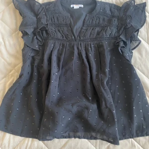 Svart blus med volang från Gina Tricot - Supersöt svart blus från Gina Tricot med volangiga ärmar och diskret prickigt mönster. Blusen har v-ringning och fina rynkade detaljer upptill. Perfekt för dig som gillar en luftig och trendig look.