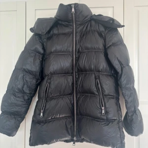 Svart pufferjacka från Massimo Dutti - Svart pufferjacka från Massimo Dutti med hög krage och avtagbar huva. Jackan har två stora fickor med dragkedja framtill och quiltad design. Perfekt för kalla dagar och har en snygg, lite glansig finish.