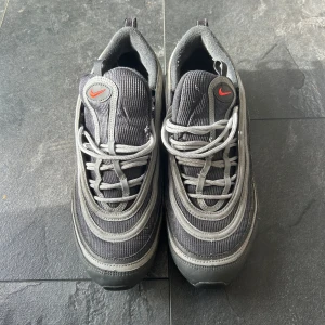 Nike Air Max 97  - Säljer ett par Nike Air Max 97 i grått och silver med röda detaljer. Skorna har den klassiska vågiga designen, snörning och en synlig Air Max-sula. Ovandelen är i syntet och mesh för en sportig look. Perfekta för dig som gillar streetwear och sneakers med attityd.