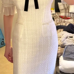 Vit elegant klänning med rosett - Supersnygg vit klänning i polyester med halvärmade ärmar och volangdetaljer. Klänningen har en svart rosett framtill, pärldetaljer vid fickorna och ett diskret rutmönster i tyget. Perfekt för dig som vill sticka ut med en chic och klassisk look.