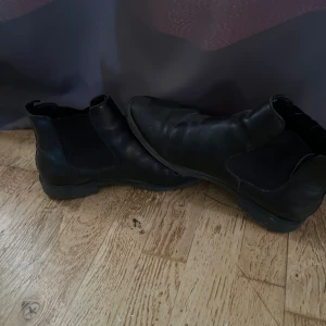 Svarta chelsea boots i skinn - Säljer ett par klassiska svarta chelsea boots i skinn med elastiska paneler på sidorna och låg klack. Skorna har rund tå och platt sula, perfekt för en stilren look. Passar till både jeans och kostymbyxor.