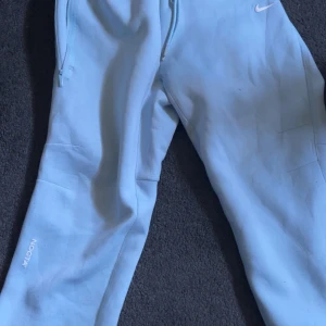 Ljusblå NOCTA x Nike mjukisbyxor - Snygga ljusblå mjukisbyxor från Nike NOCTA med dragkedjor vid fickorna och broderad logga på benet. Perfekta för en chill och sportig look. Sköna och stilrena med elastisk midja.