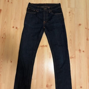Mörkblå jeans från Nudie Jeans, 30/32 - Snygga mörkblå jeans från Nudie Jeans i klassisk rak modell. Jeansen har orange kontrastsömmar, fem fickor och är tillverkade i slitstarkt denim. Perfekta för dig som gillar stilrena och tidlösa jeans med en clean look. Obs sista bilden visar på en liten missfärgning på byxorna annars perfekt skick