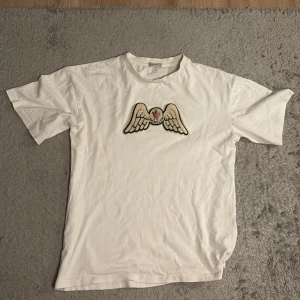 Vit Moncler t-shirt med vingar - Vit t-shirt från Moncler med broderad ving-detalj och logga på bröstet samt stor svart text 'Angels' på ryggen. Klassisk rund hals och korta ärmar. Tillverkad i mjuk bomull för en skön känsla och clean look.