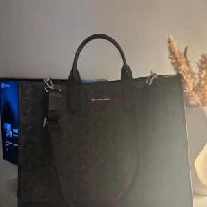 Svart axelväska från Michael Kors - Michael Kors Greyson Large Logo Briefcas Perfekt för jobb eller när man ska åka iväg Mått: Höjd: 35cm, längd: 42cm, bredd: 17cm  Färg: Svart/grå med MK-monogrammönster. Använd 1 gång Nypris ca 3000 Material: Coated canvas med läderdetaljer. Har både handtag och en avtagbar axelrem. Silverfärgade metalldetaljer.