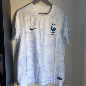 Frankrike fotbollströja Nike vit/blå - Säljer en officiell Frankrike fotbollströja från Nike i vitt med ljusblått mönster. Tröjan har korta ärmar, broderad tupp-logga och två stjärnor på bröstet. Materialet är lätt och andas, perfekt för match eller träning. Pris går att diskuteras 