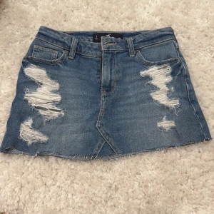 Blå jeanskjol med slitningar från Hollister - Snygg blå jeanskjol från Hollister med slitna detaljer och rå kant nertill. Klassisk femficksmodell med bälteshällor och coola slitningar framtill. Perfekt för dig som gillar en avslappnad och trendig stil.