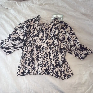 Beige och svart blus med mönster - Säljer en snygg beige blus med svart abstrakt mönster. Blusen har trekvartsärm, lätt rynkad struktur och liten volang vid halsen. Perfekt för dig som vill ha något unikt och trendigt i garderoben. Storlek L men väldigt liten i storlek skulle säga en S