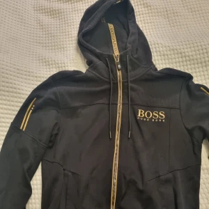 Svart hoodie från Hugo Boss - Svart hoodie från Hugo Boss med guldiga detaljer och logga på bröstet. Fast pris 325 kr. Inga byten. Se även mina andra annonser och skapa bundles.