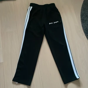 Palm Angels svarta mjukisbyxor med vita ränder - Svarta mjukisbyxor från Palm Angels med vita sidoremsor och logga tryckt på framsidan. Byxorna har elastisk midja och dragkedja vid benslut. Perfekta för en chill och sportig look.