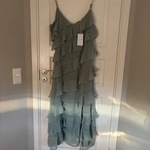 Grön volangklänning från VILA - Supersnygg långklänning från VILA i ljusgrönt med tunna axelband och flera lager volanger. Klänningen har v-ringning och en slits på sidan för extra edge. Perfekt för dig som vill sticka ut med en flowy och trendig look. Helt ny ovanvänd säljer pågrund av fel storlek. Strl 44. 