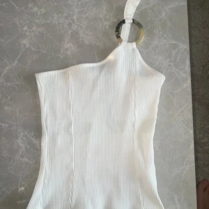 Vit ribbad one shoulder topp NA-KD - Säljer en vit ribbad one shoulder topp från NA-KD i storlek S. Toppen har en bred axelrem med en cool ringdetalj i plast och är figurnära. Perfekt för dig som gillar trendiga och stilrena plagg med det lilla extra.