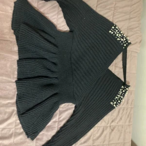 Svart stickad tröja med pärldetaljer - Supersnygg svart stickad tröja med volang nertill och långa ärmar. Tröjan har v-ringning och coola pärldetaljer på axlarna som ger en extra touch. Perfekt för dig som vill sticka ut lite extra i höst och vinter!