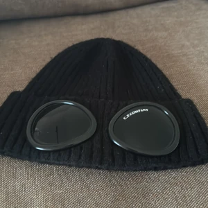 Svart C.P. Company Goggle mössa - Säljer en svart ribbstickad Goggle mössa från C.P. Company med ikoniska svarta glasögonlinser på uppviket. Mössan är tillverkad i mjuk ull och har en clean, urban look som sticker ut. Perfekt för dig som vill ha streetwear-vibe och gillar detaljer.