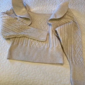 Beige stickad tröja med volangkrage - Superfin beige stickad tröja med snygg volangkrage och kabelstickat mönster. Tröjan har lång ärm och ribbade muddar vid ärmslut och nederkant. Perfekt för dig som vill ha en mysig och trendig look.