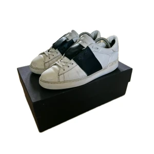 Valentino Garavani Open sneakers vit/svart - Snygga Valentino Garavani Open sneakers i vitt skinn med bred svart läderpanel över mitten. Klassisk rund tå, platt sula och vita snören. Ikoniska nitar baktill och diskret logga på plösen. Perfekt för dig som gillar stilrena och lyxiga sneakers. Dock inte orginal sula