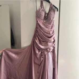 Balklänning ( rygglös)  - Säljer en elegant ljuslila långklänning med v-ringning och pärlbroderier framtill. Klänningen har draperad kjol, halterneck och glittriga detaljer längs urringningen. Materialet är glansigt och ger en lyxig känsla, perfekt för bal eller fest.