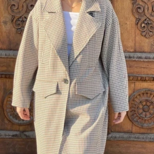 Rutig lång kappa med stora slag - Snygg rutig kappa i beige och brunt med stora slag och en klassisk krage. Kappan har en enkel knappstängning framtill och två rejäla fickor. Fodret är ljusbeige och materialet känns mjukt och lite ulligt. Perfekt för dig som gillar retrovibbar och vill sticka ut.