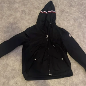 Svart vindjacka med huva från Moncler - Svart vindjacka från Moncler med huva och dragkedja framtill. Jackan har Moncler-logga på ärmen och vita samt röda ränder på huvan. Två fickor med tryckknappar och stilren design. Perfekt för dig som vill ha en snygg och sportig look.