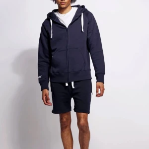 Mörkblå hoodie från Russelruss - Mörkblå hoodie med vit dragsko från Russelruss. Klassisk zip-up modell med huva, känguruficka och ribbade muddar. Perfekt för chill dagar eller när du vill ha en enkel och snygg look. Broderad logga på ärmen ger en extra detalj. PRISET KAN DISKUTERAS 